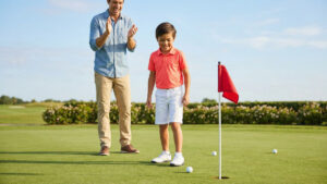 A young boy smiling on a putting green beside a red flag as his father claps in celebration, on a bright, open golf course on a sunny day.