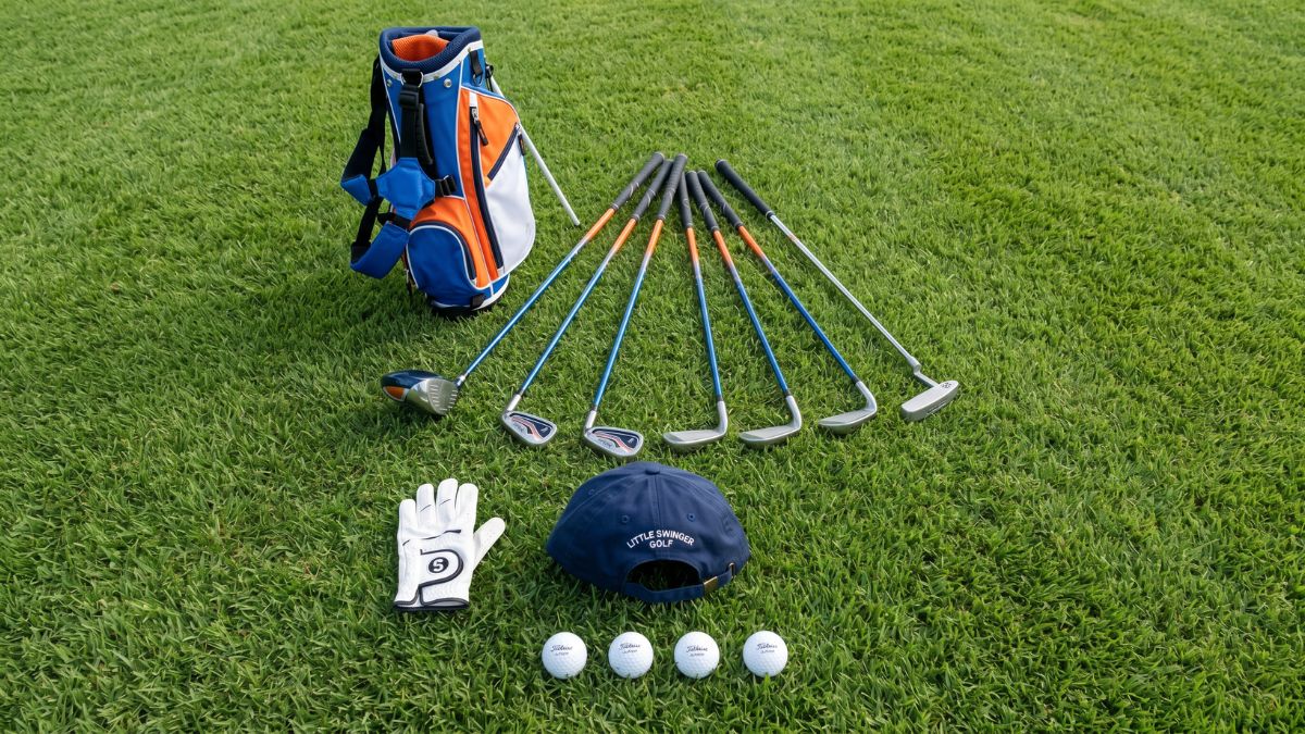 A complete junior golf starter kit laid out on short green grass, featuring a blue, orange, and white junior stand bag alongside a fanned set of junior golf clubs, a white golf glove, a navy cap, and four golf balls.