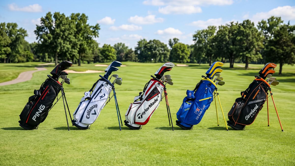 Five junior golf stand bags with full club sets from Ping, Callaway, TaylorMade, US Kids Golf, and Cleveland Golf lined up side by side on a golf course fairway against a blue sky, representing a junior golf brand comparison and review.